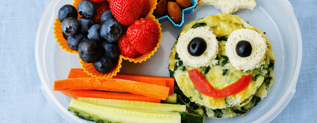 A lunch box containing fruit, vegetables and a quiche with a smiley face on it