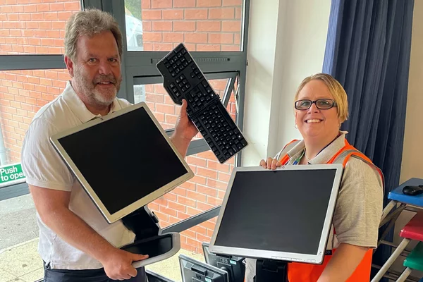 A man and a woman standing in a classroom and holding computer monitors that have been donated through the Digital Inclusion Scheme
