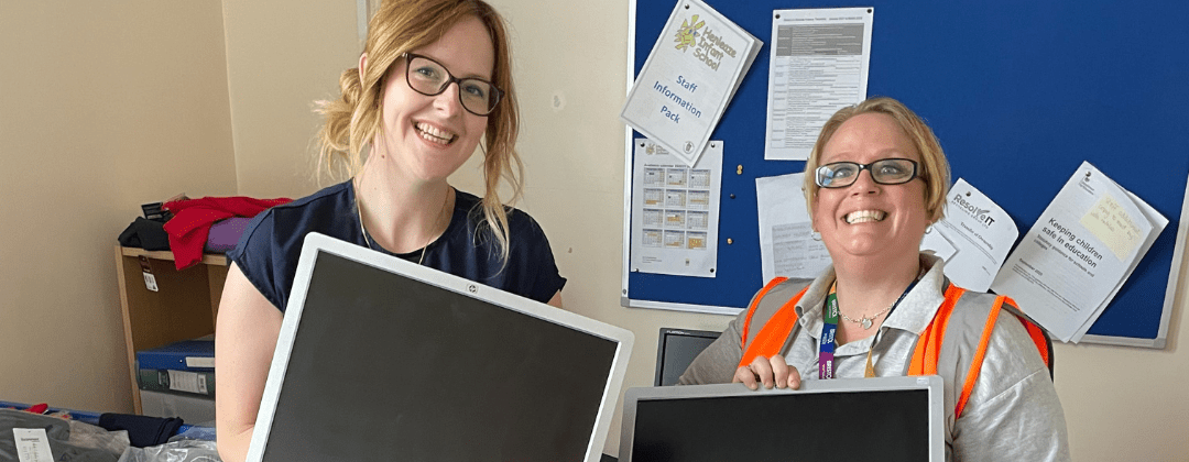 Two women standing in a classroom holding computer monitors that have been donated through the digital inclusion scheme.