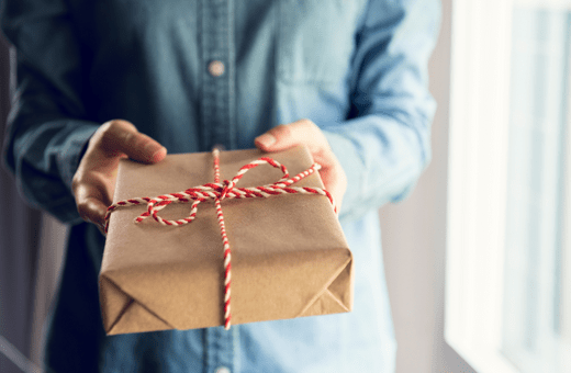 A person whose face cannot be seen is holding a present wrapped in brown paper and tied with striped string