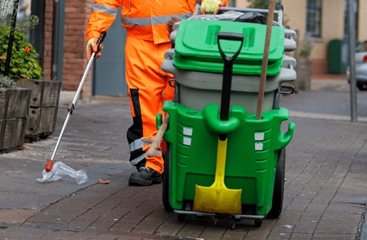 A Bristol Waste street cleaner picks up some plastic litter from the pavement using a litter picker. They are pushing a wheeled bin card with a shovel and broom attached to it.