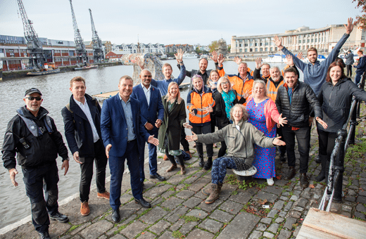 A group of Bristol Waste, Bristol City Council and Hubbub people pose in front of Bristol Harbour. The artist of the wave and her team are also there. In the background the wave sculpture is floating on the water