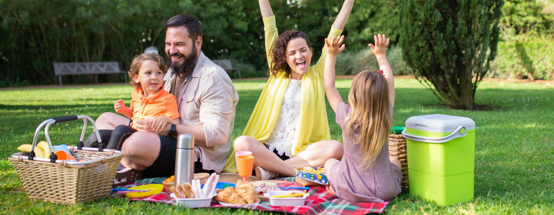 A family of four enjoy a low waste picnic in a park. They have reusable plates and cutlery, a picnic basket and a cool box on a blanket.
