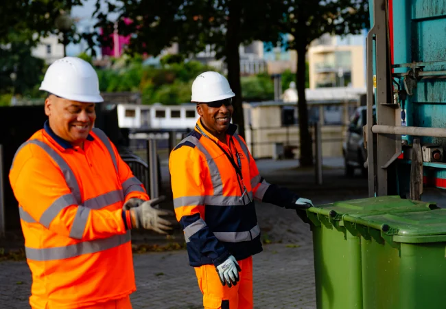 Two men in hi visibility clothing and hard hats pushing wheelie bins