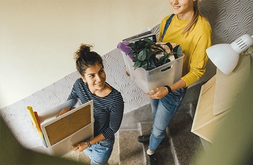 two students walk down stairs carrying boxes