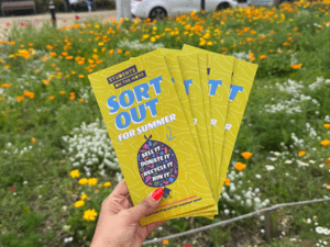 A perfectly manicured hand holds up a fan of "Sort Out For Summer" students on the move leaflets. In the background is a meadow of wildflowers