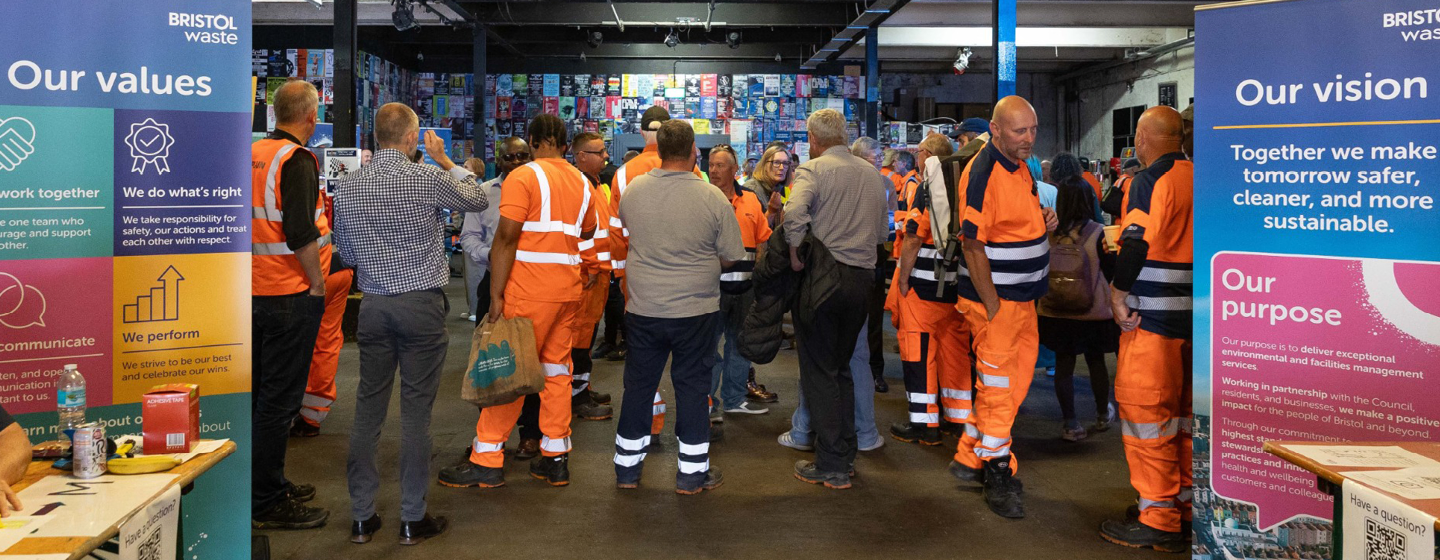 A group of people, many in high-visibility orange work uniforms, gather in a warehouse between two Bristol Waste banners.