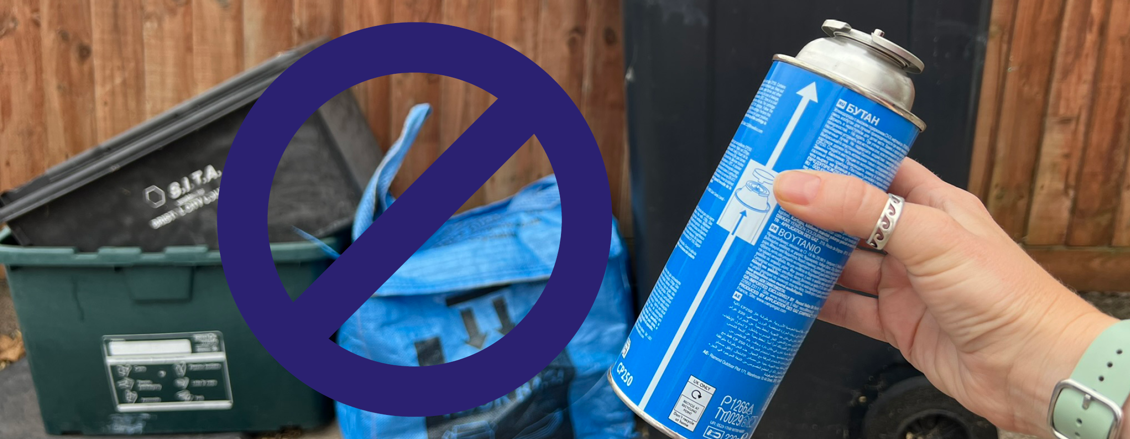 A hand holds up a used gas bottle. In the background are a set of bins and recycling containers. There is a clear "no" sign over the bins, to say that the gas canister should not go in the bins or recycling containers.