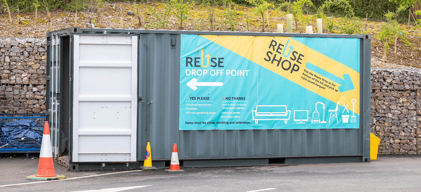 The Reuse Shop drop off point, a container with a bright sign on the outside with details of what can be dropped off. Behind is a stone wall, trees and wildflowers