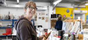 A woman with brown hair and glasses smiles at the camera as she browses ornaments in the Reuse shop