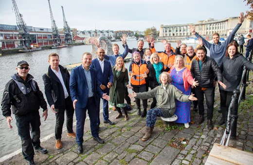 A group of Bristol Waste, Bristol City Council and Hubbub people pose in front of Bristol Harbour. The artist of the wave and her team are also there. In the background the wave sculpture is floating on the water
