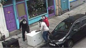 Three people creating a fly-tip, whose faces are blurred, are ditching a piece of unwanted furniture on a pavement