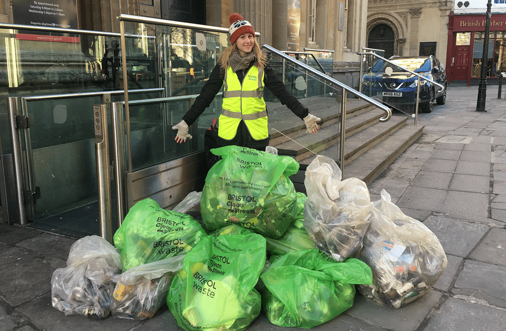 A woman stands behind a pile of litter bags with her arms outstretched