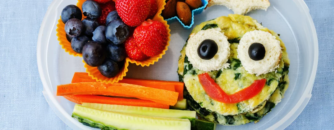 A lunch box containing fruit, vegetables and a quiche with a smiley face on it