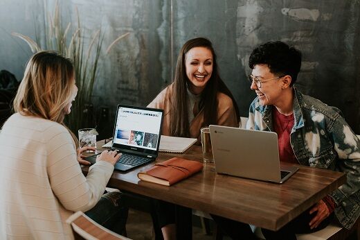 three students sitting around a table with laptops laughing