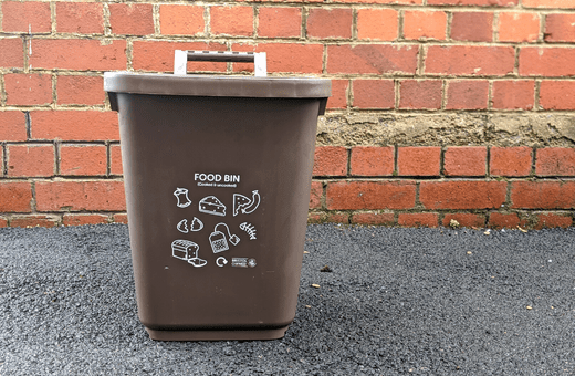 A brown food waste bin on the ground in front of a redbrick wall.