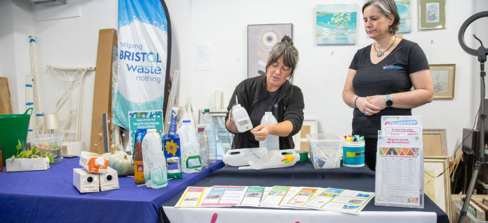 Two women from Children's Scrapstore are standing behind a table demonstrating how to turn a milk carton into a bird feeder.