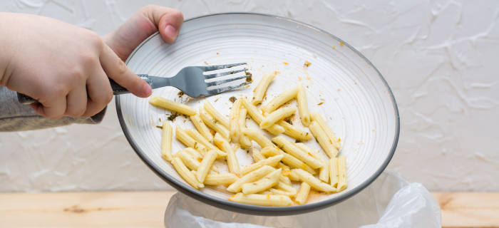 A plate of pasta is being scraped into a food waste bin by an unseen person.