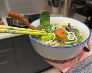 One of the low-waste recipes. A bowl of vegetable noodles is held in front of the camera. There are chop sticks and a spoon sticking out of the bowl and in the background are kitchen cabinets