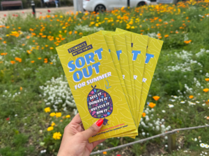 A perfectly manicured hand holds up a fan of "Sort Out For Summer" students on the move leaflets. In the background is a meadow of wildflowers
