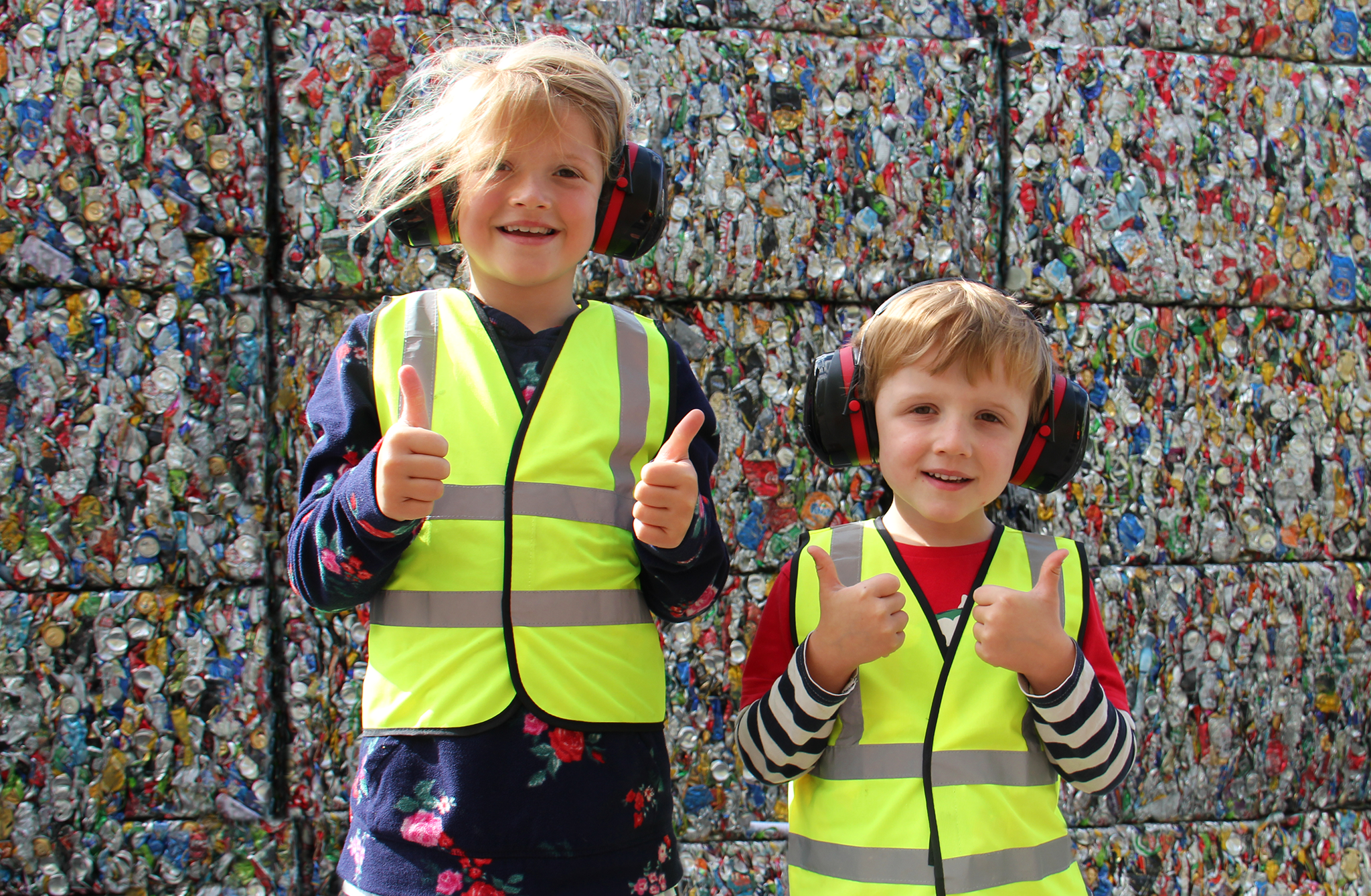 Two small children in hi-viz vests and ear defenders standing in front of bales of compacted cans and giving thumbs up