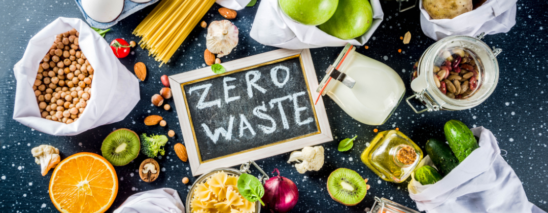 The words 'zero waste' are written on a chalkboard and surrounded by reusable containers of dried pasta and fresh fruits.
