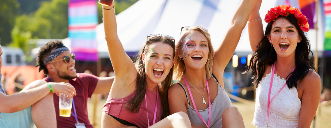 A group of friends sitting on the grass at a music festival. Three women are smiling at the camera and raising their arms in the air in celebration.