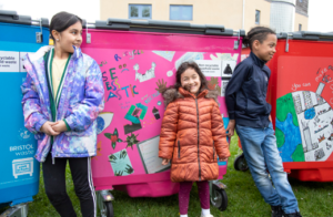 Young artists standing next to their designs on blue, pink and red bins 