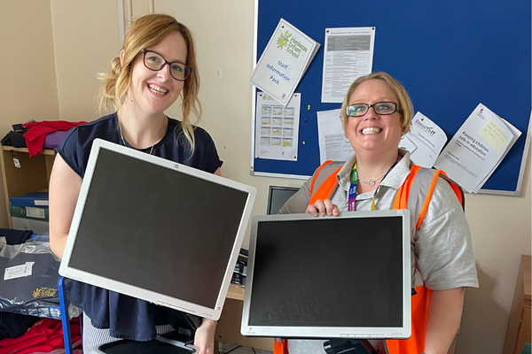 Two women standing in a classroom holding computer monitors that have been donated through the digital inclusion scheme.