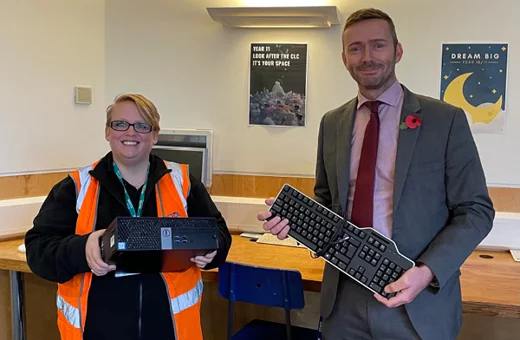A man and a women smiling holding a desktop computer and keyboard inside a classroom