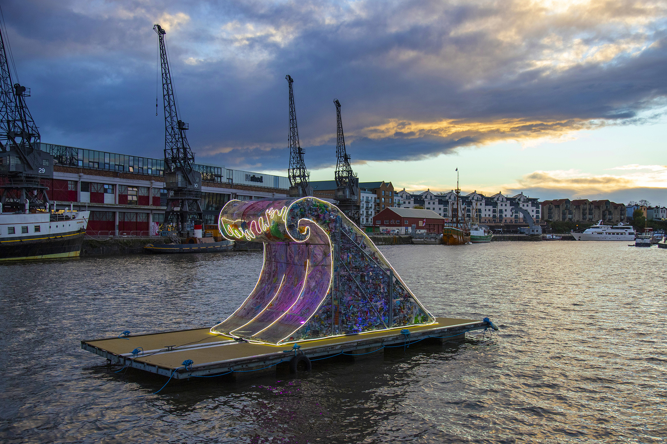 A giant wave sculpture made from rubbish sits on a floating pontoon on Bristol harbour. In the background are the iconic Bristol cranes outside of the M Shed. The clouds in the sky are grey and moody and the sculpture is lit up with strings of coloured lights.
