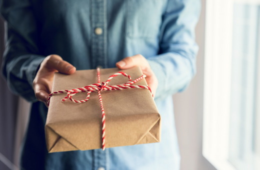 A person whose face cannot be seen is holding a present wrapped in brown paper and tied with striped string
