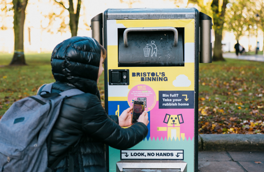 A young person in black puffer jacket and backpack uses their smartphone to scan the QR code on a brightly coloured Bristol bin