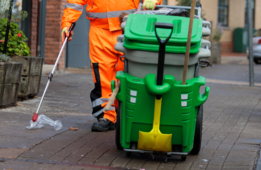 A Bristol Waste street cleaner picks up some plastic litter from the pavement using a litter picker. They are pushing a wheeled bin card with a shovel and broom attached to it.