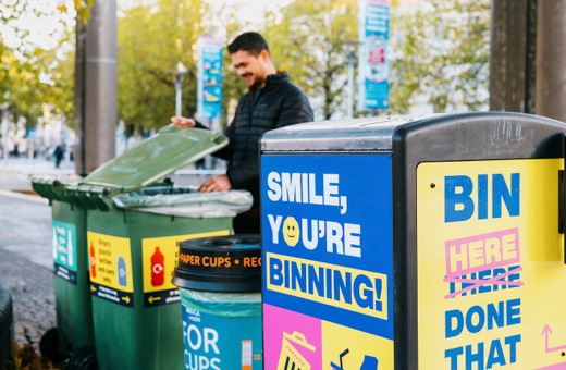 A smiling person lifts the lid of a Bristol's Binning glass recycling bin on Bristol city centre. They look like they're just recycled something in the bin