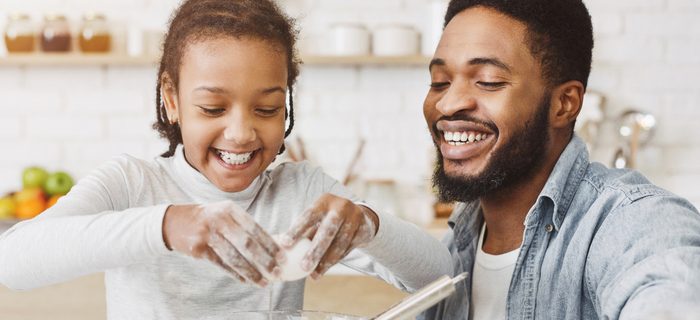A father smiles at his daughter who is cracking eggs into a bowl to make pancakes.