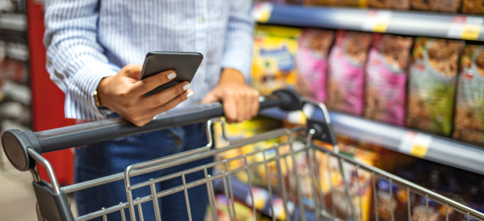 In a supermarket aisle, a person looks at a shopping list on their phone while pushing a trolley.