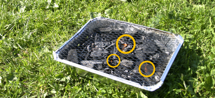A disposable barbecue that has been left on the grass. Small flames can be seen appearing among the coals. 