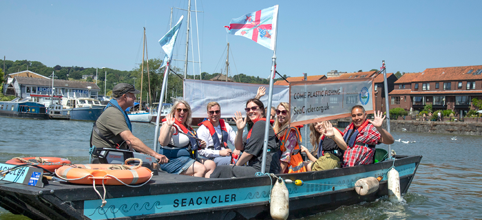 A crew of people sit on board the Sea-Cycler on the water. The Sea-Cycler is a boat owned by Sustainable Hive, perfect for water-born litter picking. The sun is shining and the people are waving at the camera. 
