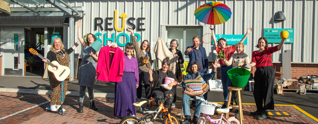 It's a sunny day with a bright blue sky. A group of people are posing for a photo outside the Reuse Shop at the Hartcliffe Way recycling centre, smiling and holding a fun variety of items in outstretched arms: a guitar, a rainbow umbrella, a pink jacket, scooters and a plant pot. They are here to promote the launch of the Waste Nothing Challenge.