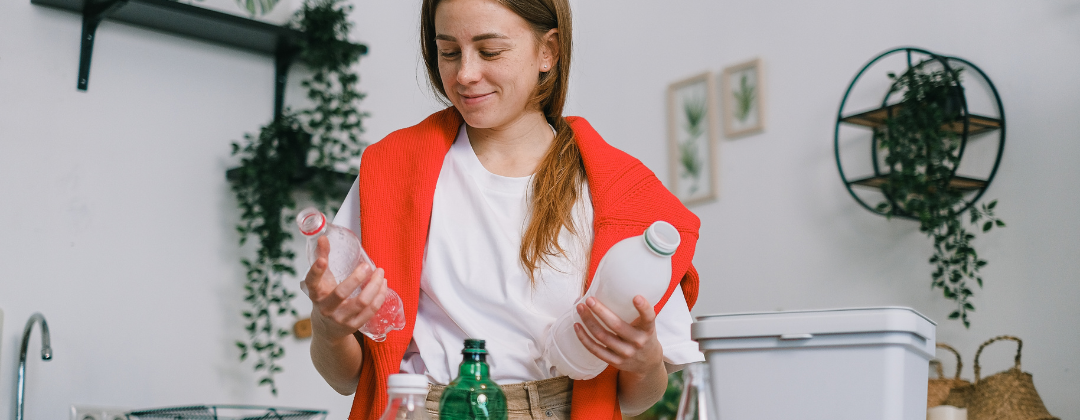 A woman, standing in her kitchen, is looking at two plastic bottles in her hands before putting them in the recycling bin.