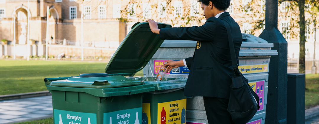 A boy in school uniform is putting an empty plastic bottle into a recycling bin near College Green