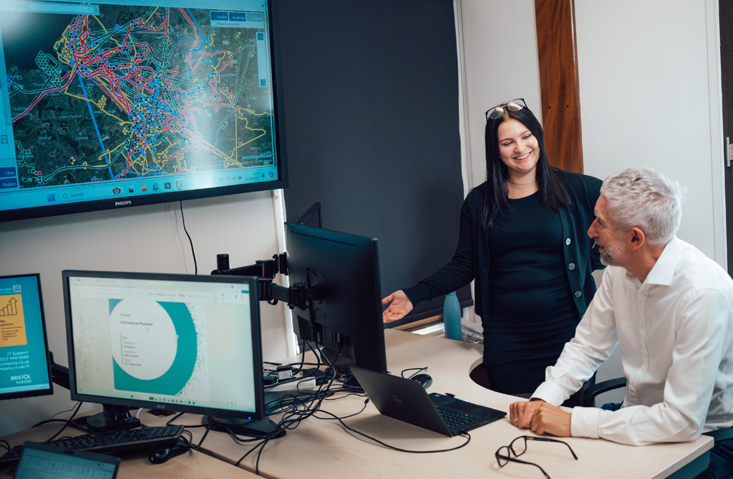 Two people are in an office. On a large screen on the wall, they are looking at a mapping system for Bristol