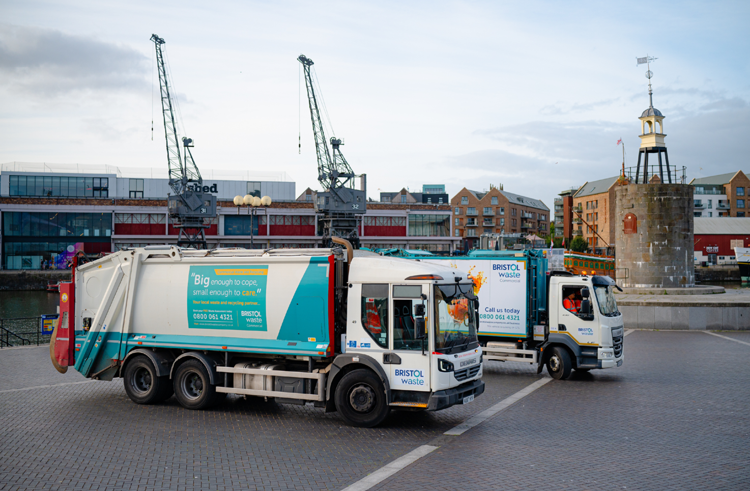 Two waste trucks are parked by Bristol Harbour. The iconic Bristol cranes are visible in the background