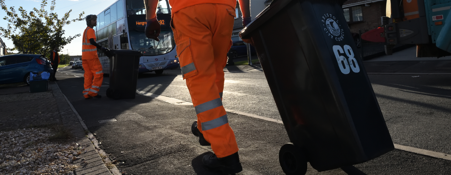 Two people in hi-vis are collecting general waste wheelie bins and emptying the contents into the Bristol Waste truck