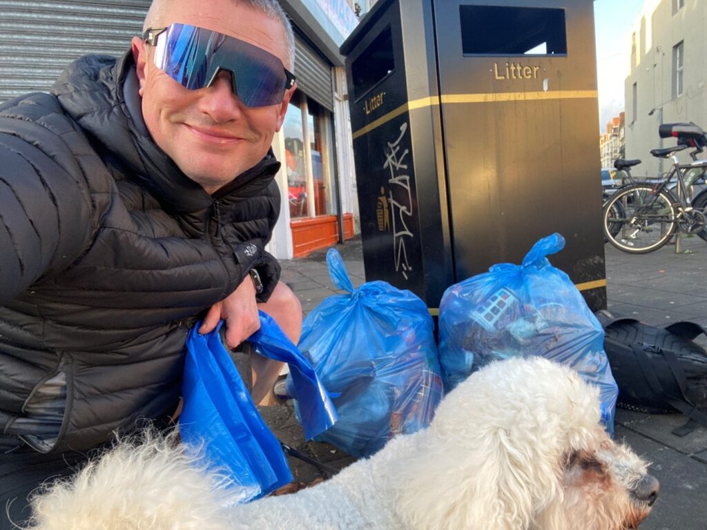 Mark Rowlands, wearing a puffer jacket and sunglasses crouches by a litter bin. There are bags of litter he's collected in the background and a fluffy, white dog in the foreground
