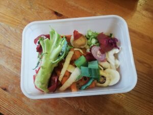 One of the low-waste recipes: A Tupperware tub sits on a wooden counter. The tub contains vegetable peelings, a broccoli stem, ends of various root veg and other veggie offcuts