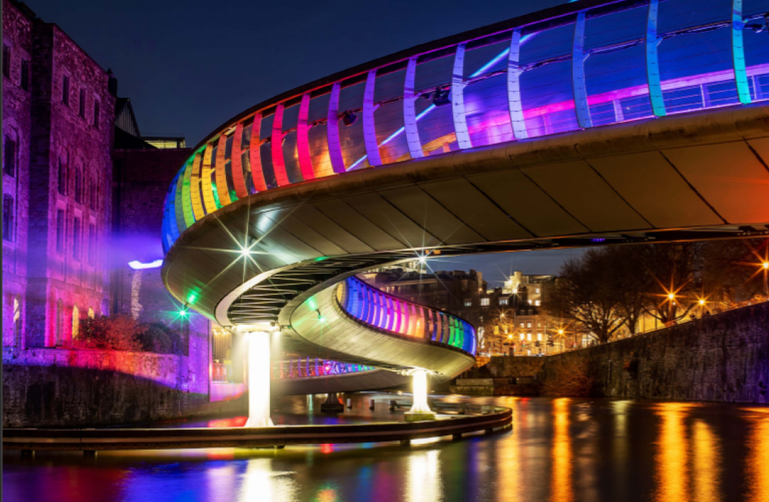 Castle Bridge in Bristol at dusk. The bridge is lit up by a rainbow of lights which reflect in the water below.