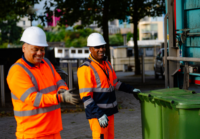 Two men in hi-vis clothing and hard hats next to wheelie bins