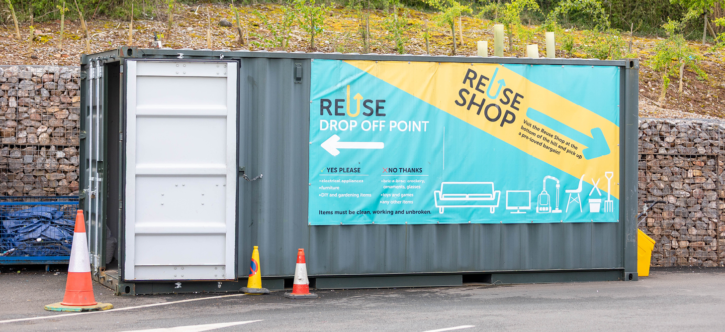 The Reuse Shop drop off point, a container with a bright sign on the outside with details of what can be dropped off. Behind is a stone wall, trees and wildflowers